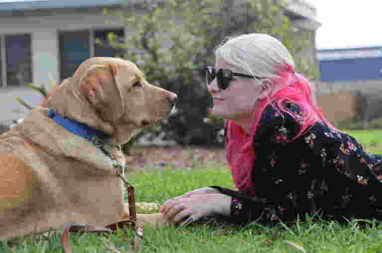 A young girl laying on grass, holding her Guide Dog's paws. Her yellow Labrador Guide Dog and her are facing each other.