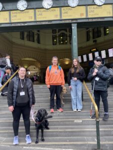 Four camp attendees stand under the clocks on the Flinders St Station steps. Two attendees have a Guide Dog and one holds a white cane.