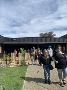 A group of Clients walking along a gravel path in our sensory garden, holding their white canes. To the left is a patch of grass with a fenced off pond in the background.