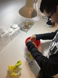 Matt stands at a white kitchen bench pouring 2-minute noodles and flavouring into a red bowl. A desk lamp is positioned over the bowl illuminating his view.