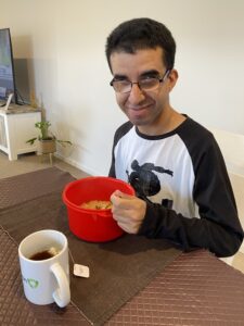 Matt sits at a dining table with a cup of tea and a red bowl full of noodles he made for himself using the help of his Occupational Therapist. He looks very proud.