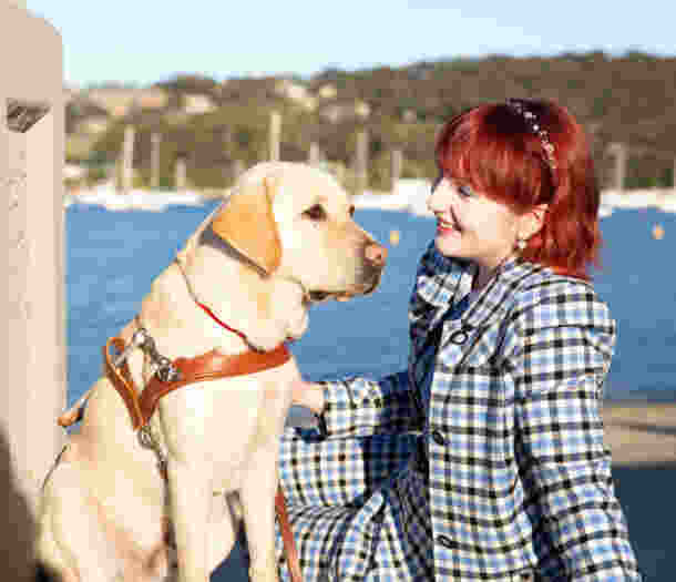 A Client smiling at her yellow Labrador Guide Dog in harness, as they sit together on a pier.