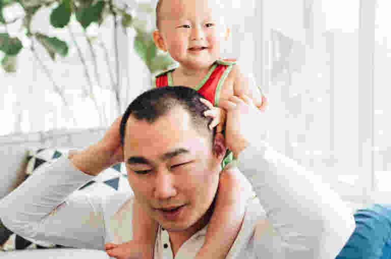 A young Asian blind man with his son at home. His son sits on his shoulders as he holds him around the waist. Big windows and greenery is in the background.