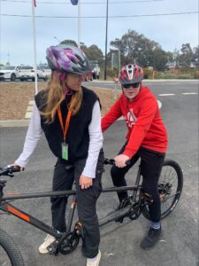 Two people wearing helmets sit on a tandem bicycle outside the Guide Dogs Kew Campus.