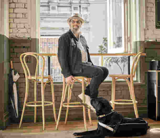 A person sitting on a stool at a cafe, with their black Guide Dog in harness at their feet.