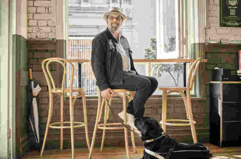 A person sitting on a stool at a cafe, with their black Guide Dog in harness at their feet.