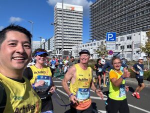 Four Berlin Marathon runners look into the camera during their run. Buildings and other runners can be seen behind them.