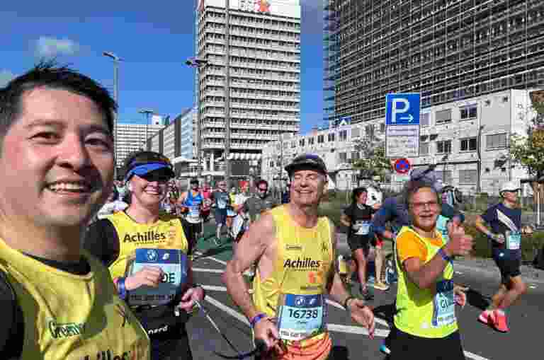 Four Berlin Marathon runners look into the camera during their run. Buildings and other runners can be seen behind them.