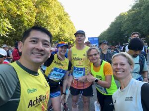 Five people smiling at the camera stand in a group on the Berlin Marathon track. Trees line the background on both sides of them.