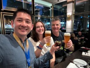 Three people smiling into the camera sit at a bar after completing the Berlin Marathon. Two of them hold glasses of beer.