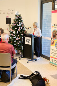 GDV team member Susannah stands at a lectern reading a poem. She stands in front of a large Christmas tree decorated in origami ornaments.