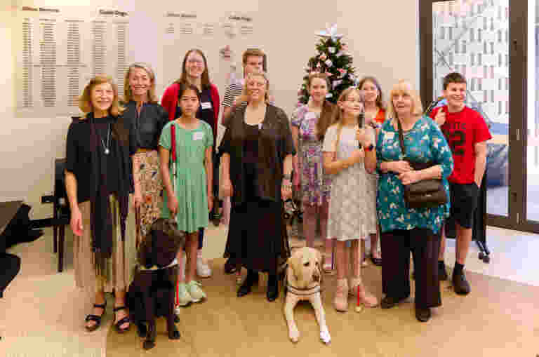 A choir of six children and three adults with low vision or blindness, accompanied by a yellow Guide Dog and a black Guide Dog, GDV CEO, Nicky Long and a support worker.