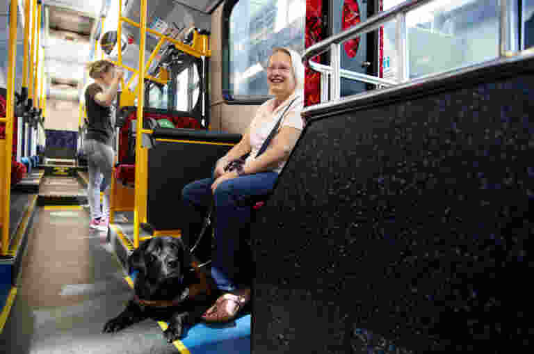 A Guide Dog Handler sits on a bus with her black Guide Dog tucked safely out of the way under her bus seat.