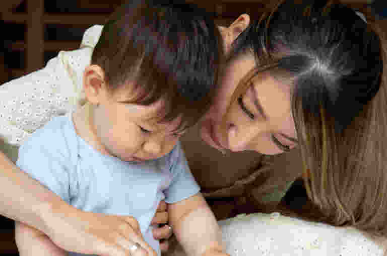 A mother guiding a child playing with red play-doh on a low table. The mother has her arm around the child and she points at the play-doh.