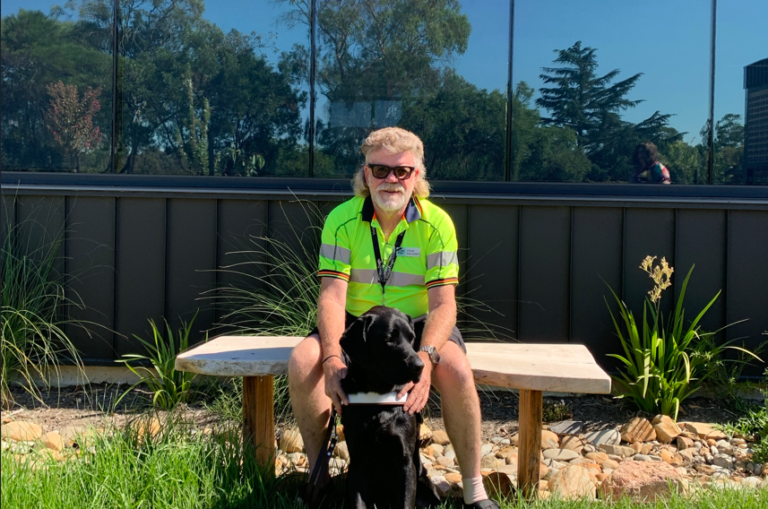 David sits at a bench outside with Lenny at his feet looking at the camera.