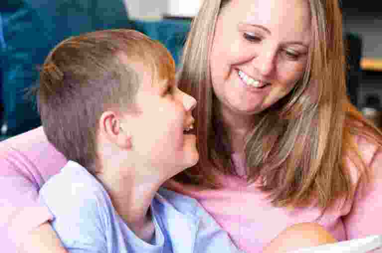 A parent with their arm wrapped around their child, holding a book. Seated in front of a couch. The parent and child are smiling at each other.
