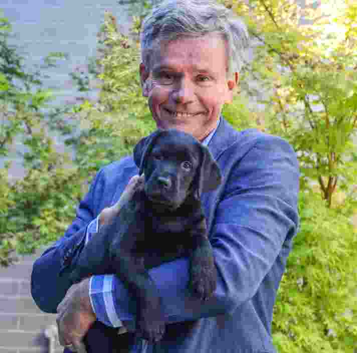 James Elder holding a black Labrador puppy in his arms, smiling at the camera.