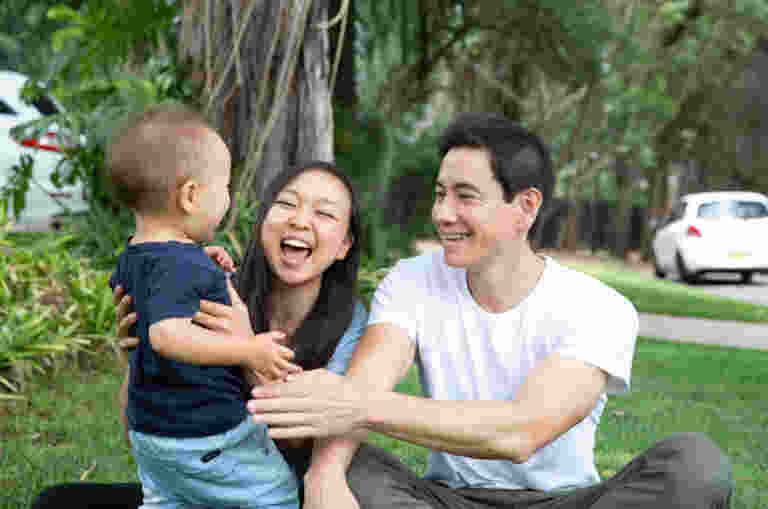 A young family with a toddler sit on grass and have a laugh.