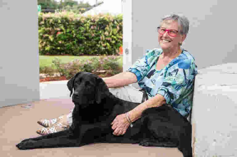 Guide Dogs Client, Sue sits on the concrete outside with her hand on black Guide Dog, Vegas.