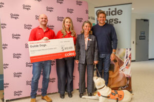 Coles Team presents a large donation check to Guide Dogs. Two Coles staff stand with Nicky Long and Curtis Stone. Cookie, a yellow Labrador wears an orange training coat in front of them. A Guide Dogs branded media wall is in the background.