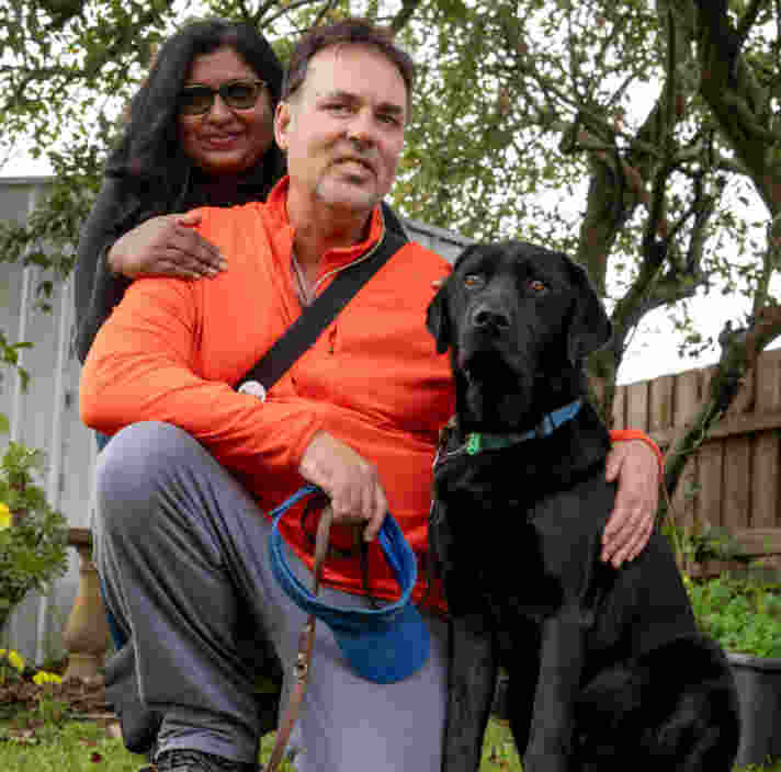 David kneels in grass with his arm around his sitting Guide Dog Fraser next to him. His wife has her hand on his shoulder crouched behind him. Both are smiling to camera.