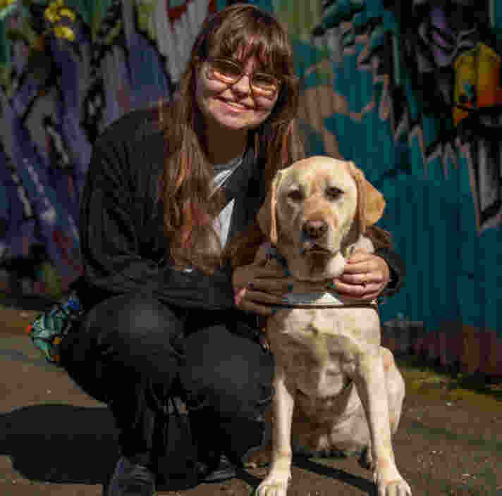 Lottie crouching down beside her sitting Guide Dog Kenita. Lottie smiles at the camera. They are sitting in front of a graffiti covered fence.