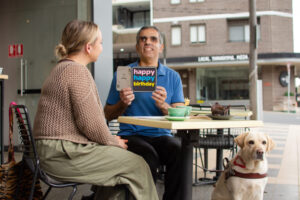 Two people at an outdoor café table, one holding a "happy happy birthday" card, with a Guide Gog beside them.