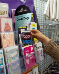 A person choosing a greeting card from a store display with a sign that reads "Hallmark Proudly supporting Guide Dogs."