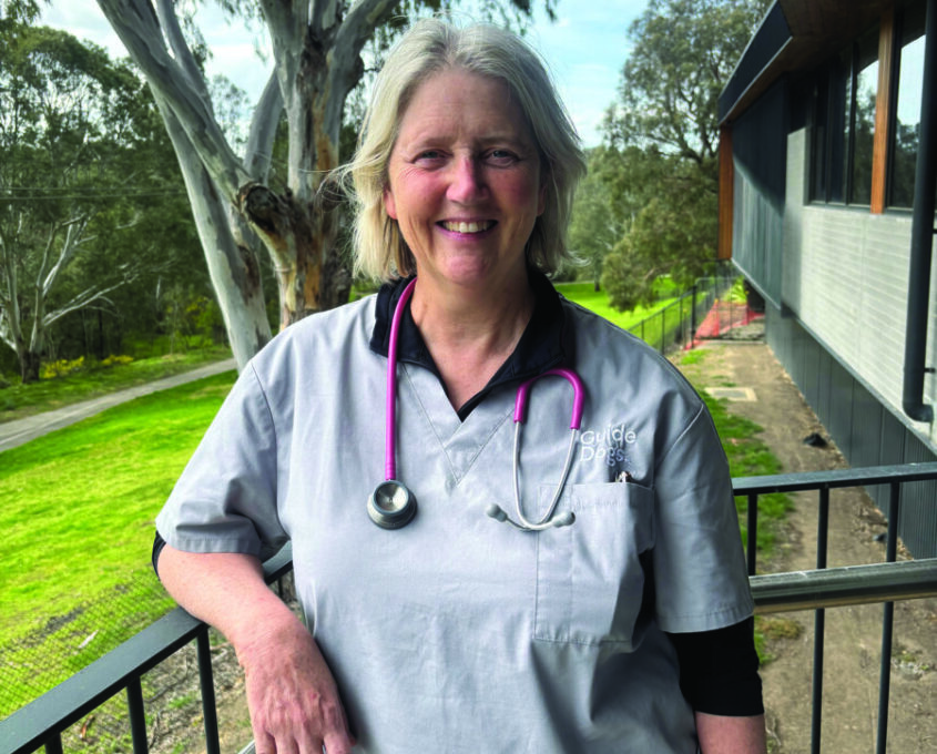 Janice smiling at camera after working in the vet clinic wearing grey Guide Dogs branded scrubs anda pink stethoscope around her neck.