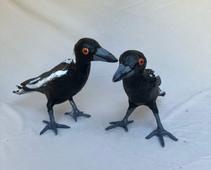 Two paper mache sculptures of black and white birds with orange eyes, standing on a coloured surface.