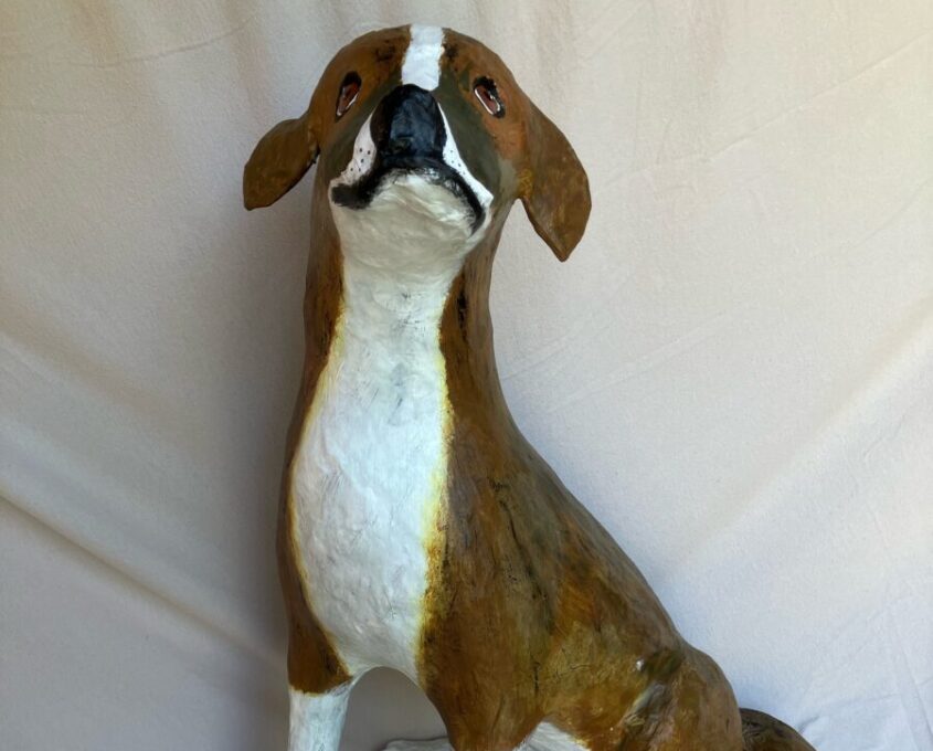 Paper mache sculpture of a dog sitting with brown fur and white belly, legs, and tail tip, head tilted upward and ears flopped back.
