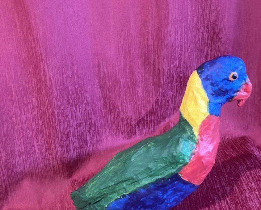 Colorful paper mache sculpture of a parrot with a blue head, yellow neck, green wings, red chest, and blue tail, set against a pink textured fabric background.