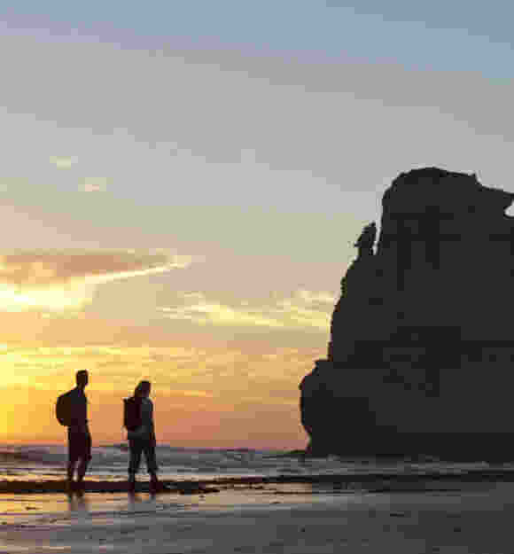 Two people with backpacks stand on a beach at sunset, facing a large rock formation in the water.