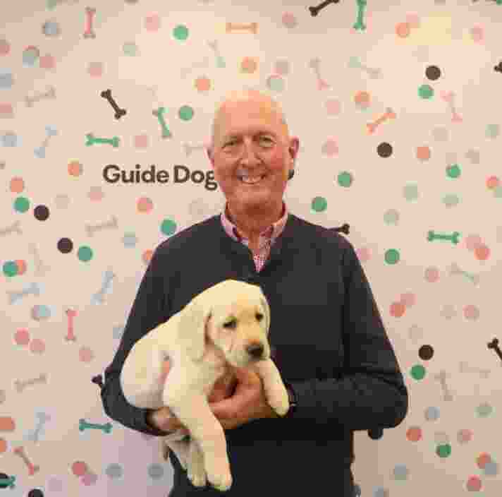 Michael Boyce holds a yellow lab puppy smiling at the camera in front of a Guide Dogs branded wall.