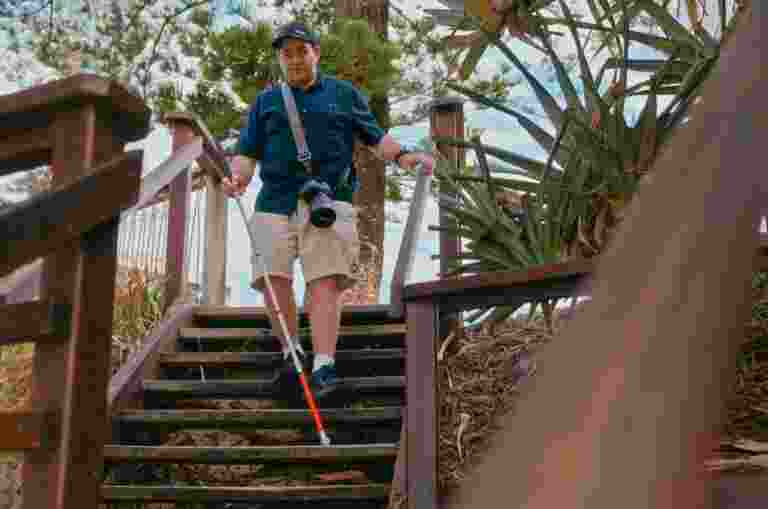 James wearing a blue shirt and beige shorts walks down a wooden outdoor staircase using a white cane. They have a camera slung over one shoulder, and the surrounding area is filled with trees and greenery.