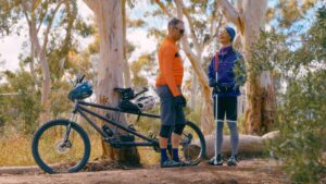 Two people stand in a wooded area with tall trees and green foliage. One wears an orange long-sleeve shirt, black shorts, and blue socks, standing beside a tandem bicycle. The other wears a purple jacket and grey pants, holding a white cane. 