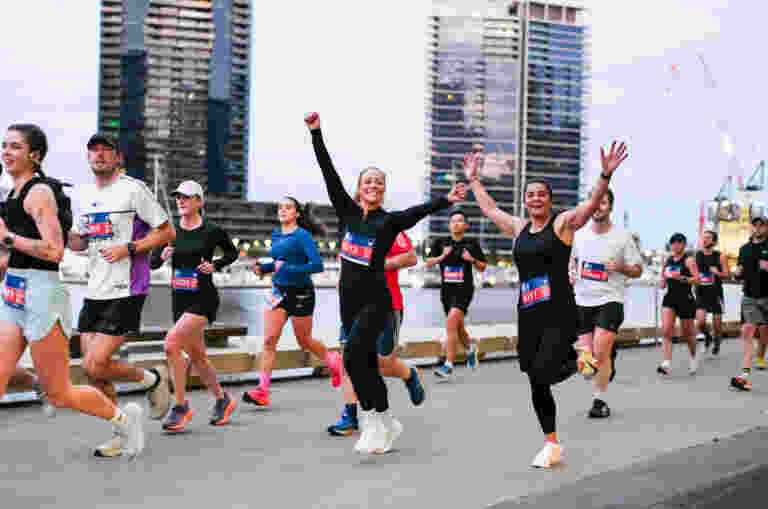 Two women in exercise gear and marathon bibs running with their arms raised in the air smiling at the camera. Other runners dispersed behind them.