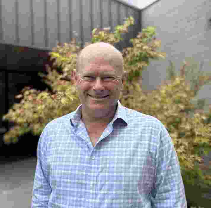 Chris McFadden wearing a light blue checkered button-up shirt stands facing the camera in an outdoor courtyard with leafy trees and modern grey buildings in the background, lit by natural daylight.
