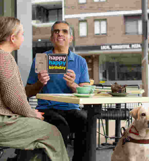 Guide Dogs client Andrew sits at a café table holding a black "happy happy birthday" card with pink, blue, and yellow text