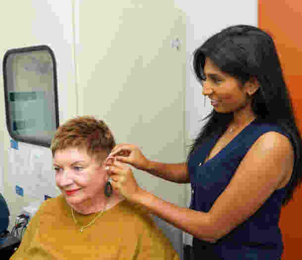 Person fitting a hearing device behind another person’s ear in a clinical room, with diagnostic equipment and cabinets visible in the background.