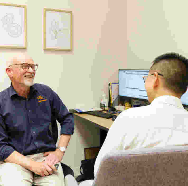 Two people seated in a small consultation room, facing each other across a desk with dual computer monitors displaying software, framed line artwork on the wall, and office chairs arranged for discussion.