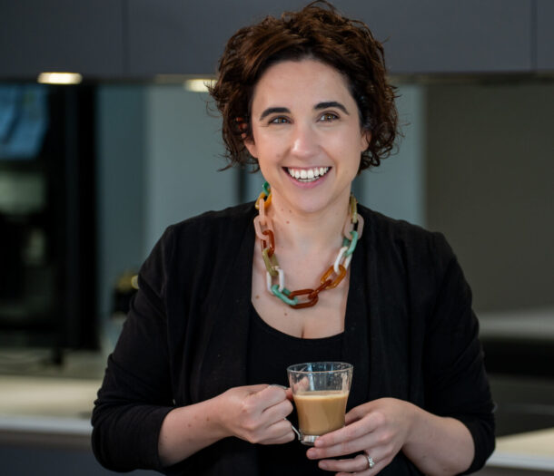 A smiling woman with short dark curly hair holds a glass mug of coffee while standing in a modern office kitchen. She is wearing a black top and cardigan with a colourful statement necklace. The background features grey cabinetry, a kitchen bench and a bowl of fruit, softly blurred.
