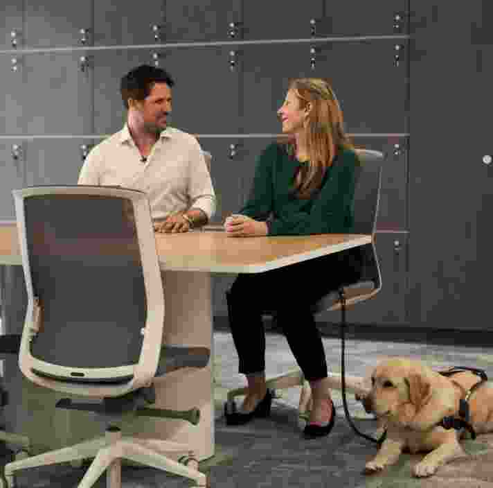 Two colleagues sit at a meeting table in a modern office, engaged in conversation. A yellow Labrador Guide Dog wearing a harness lies calmly beside one of the employees, highlighting an inclusive workplace where a Guide Dog is part of the working environment.