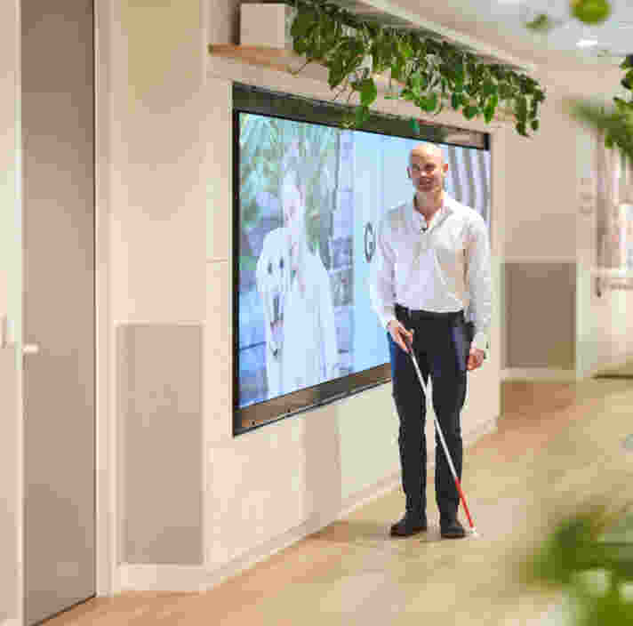 A man walks confidently through a modern office using a white cane while carrying a folder. A large digital display and indoor plants line the hallway, highlighting an accessible and inclusive workplace environment.A man walks confidently through a modern office using a white cane while carrying a folder. A large digital display and indoor plants line the hallway, highlighting an accessible and inclusive workplace environment.