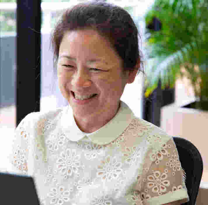 A smiling woman sits at a desk using a laptop in a bright office. She is wearing a white blouse with a floral lace pattern, with large windows and indoor plants in the background.