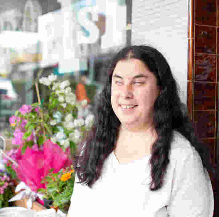 A smiling woman with long dark hair stands outside a flower shop, with colourful flowers and plants displayed beside her. She is wearing a white top and looking slightly to one side, with the shopfront in the background.
