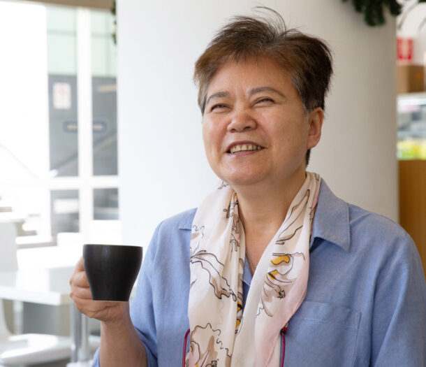 A smiling woman sits in a bright café holding a black coffee cup. She is wearing a light blue shirt and a patterned scarf, with a pair of glasses hanging around her neck. The background is softly blurred, showing tables and chairs in a light-filled indoor space.