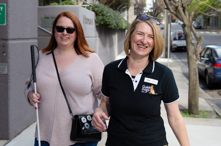 A Guide Dog staff member is guiding a Client down a footpath. They are both smiling.