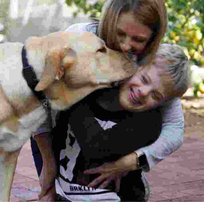 A young child is getting a kiss from a yellow labrador dog. His mother is behind them giving them a cuddle.