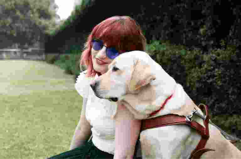 A person and a yellow Guide Dog, in harness, sitting on grass outside. The person is smiling at the camera and the Guide Dog is a profile view.